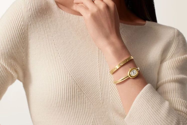 A woman in a white sweater wearing a gold Cartier bangle watch paired with a Love bracelet.